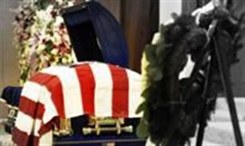A casket draped with an American flag is surrounded by flowers and a wreath in an indoor setting.