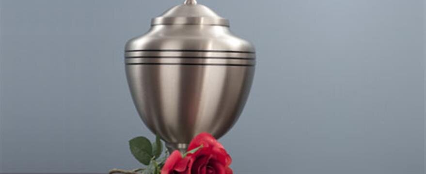A silver urn with black stripes sits on a surface, accompanied by a red rose in front of it.