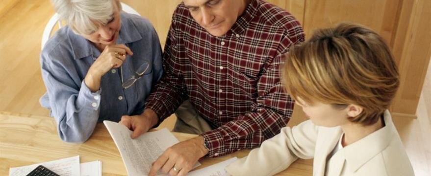 Three people sit at a table reviewing documents together, one person pointing to a page while others listen and look on.