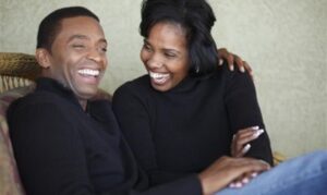 Two people sitting on a couch, smiling and laughing together, wearing black tops with relaxed postures.
