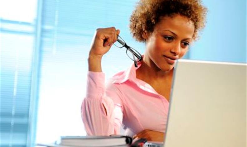 Woman in a pink shirt looks at a laptop screen while holding her glasses, with books on the table in front of her.