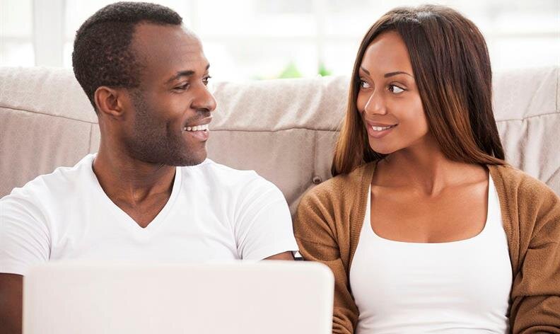 A man and woman sit on a couch, facing each other and smiling, with a laptop in front of them.