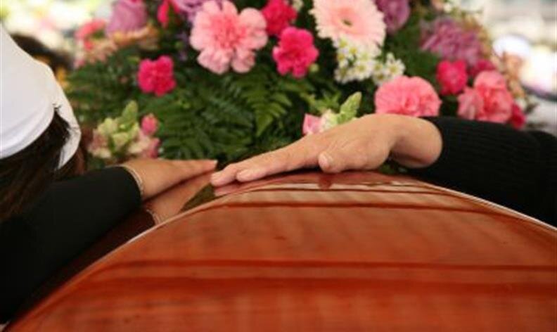 Two people rest their hands on a wooden casket with a bouquet of pink flowers in the background.