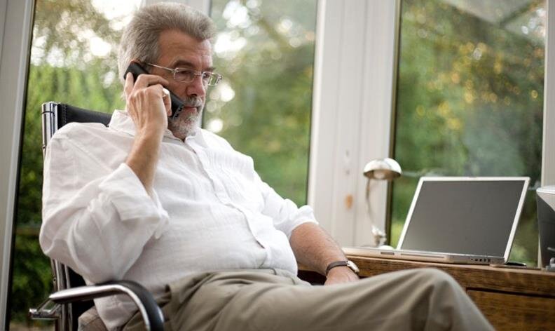 Older man in glasses sitting at a desk, talking on a phone, with a laptop open in front of him.