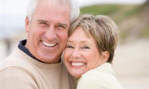 Smiling older couple embracing outdoors, both wearing light-colored clothing.