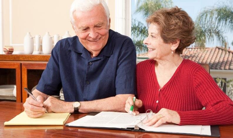 Two older adults sit at a table, smiling and working on paperwork together with a pen and highlighter.