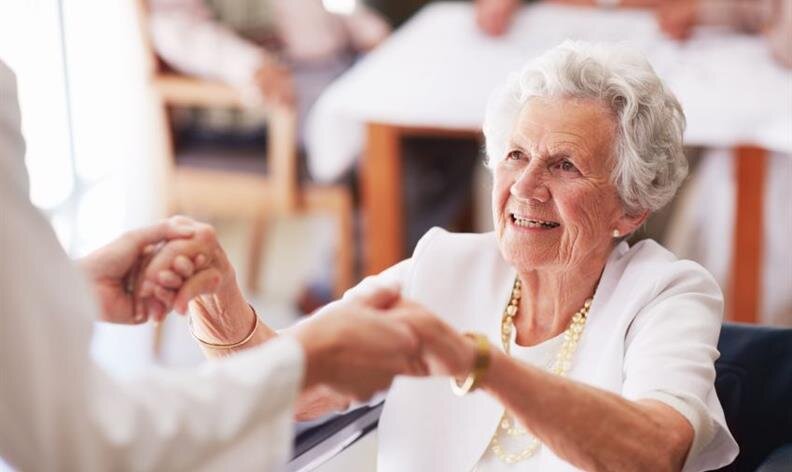 Smiling elderly woman in white outfit holding hands with another person in a bright room.