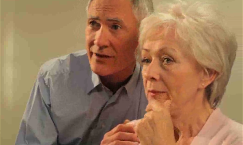 An older man and woman sit closely together, both looking ahead with concerned expressions.