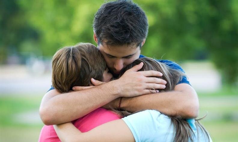 Three people stand outdoors, embracing each other closely with comforting hugs amidst a blurred green background.