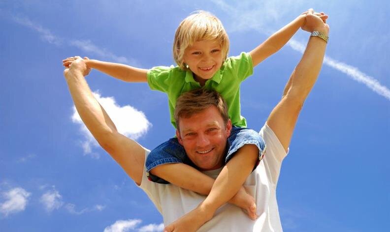 A man carries a smiling child on his shoulders outdoors under a blue sky with clouds.