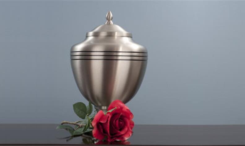 A silver cremation urn with black stripes sits on a dark surface next to a single red rose.