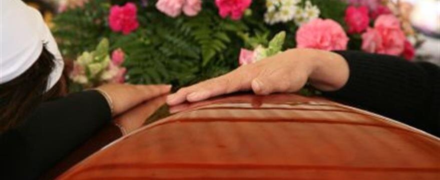 Two people rest their hands on a wooden casket adorned with pink flowers and greenery at a funeral.