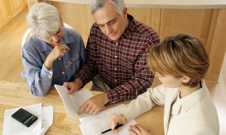 Two older adults review documents with a professional at a wooden table with papers and a calculator.